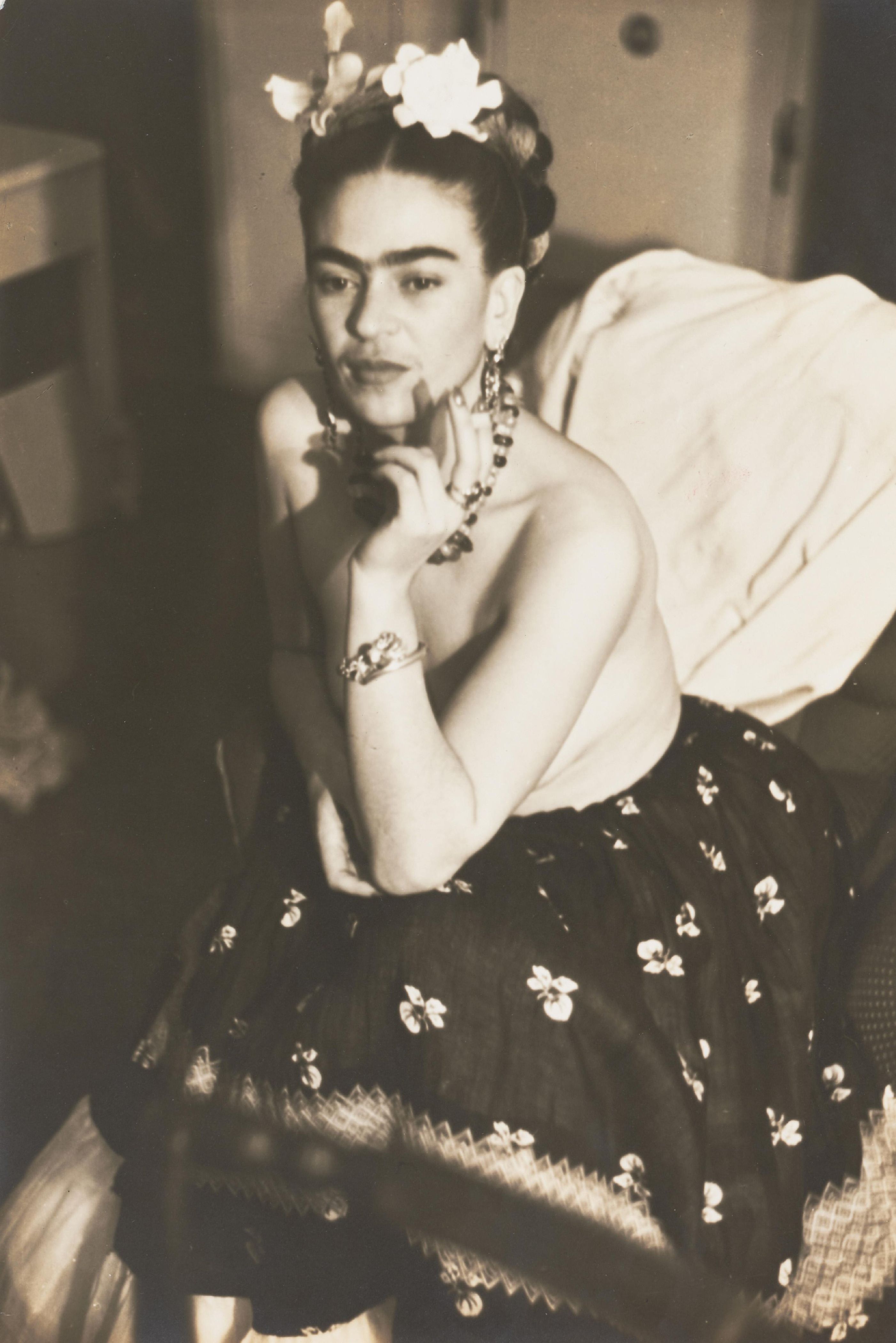 Frida Kahlo photographed by Julien Levy in 1938, posing with her arms crossed, wearing a floral headdress, ornate jewelry, and a dark floral skirt.