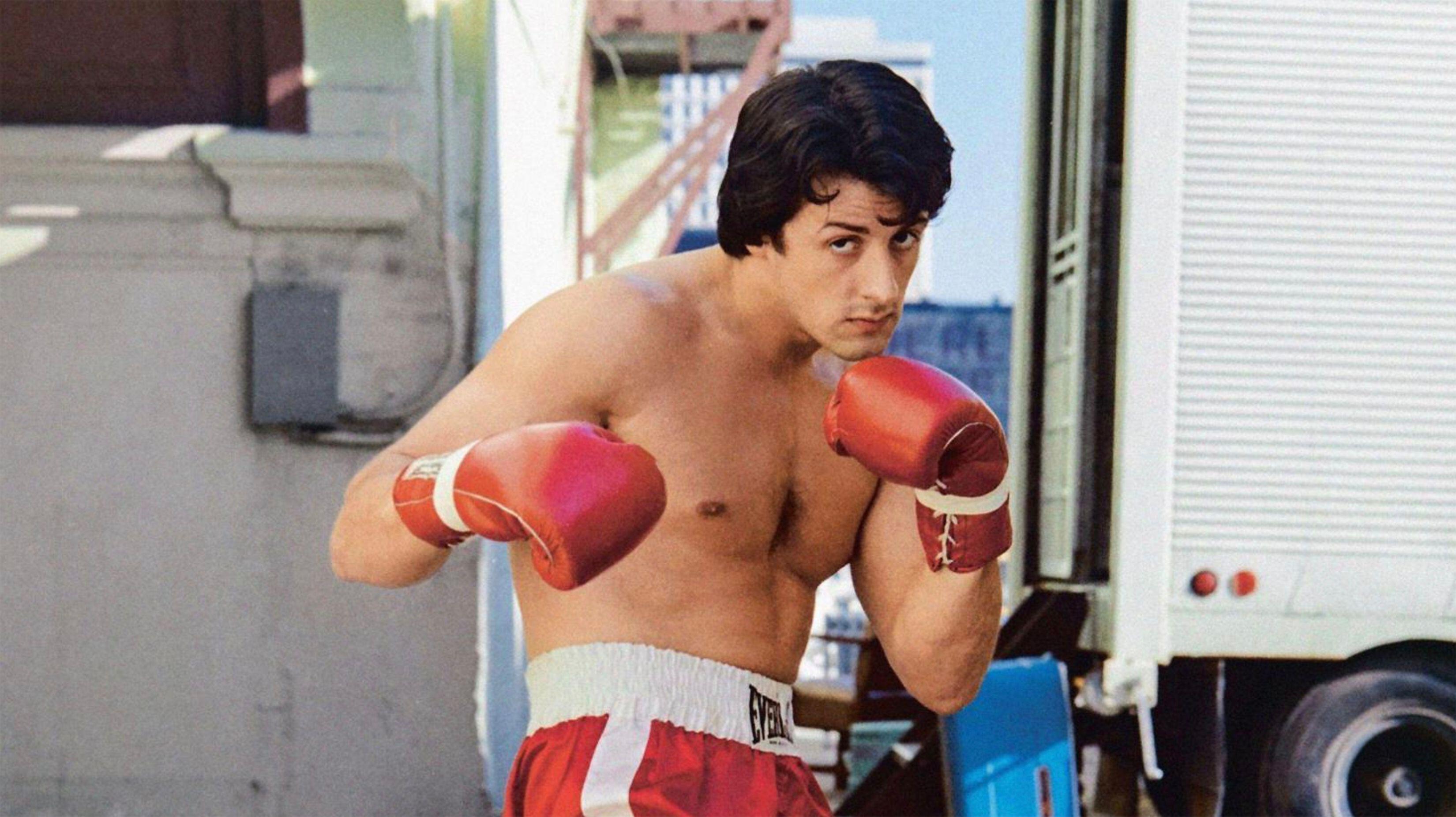 Sylvester Stallone as Rocky Balboa, shirtless and wearing red boxing gloves and shorts, in a boxing stance.