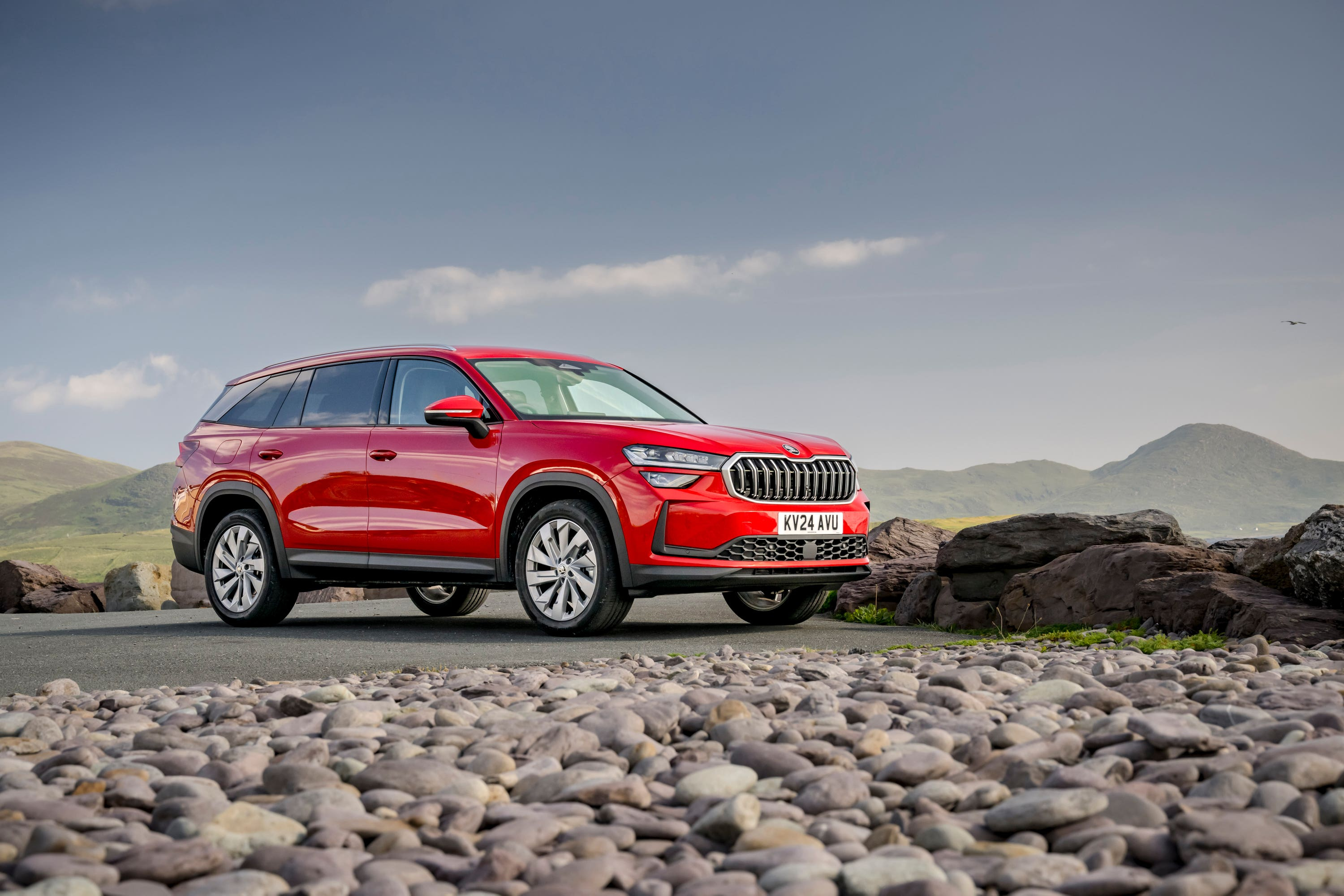 Red Skoda Kodiaq SUV parked on a paved road with a rocky foreground and green hills in the background under a blue sky.