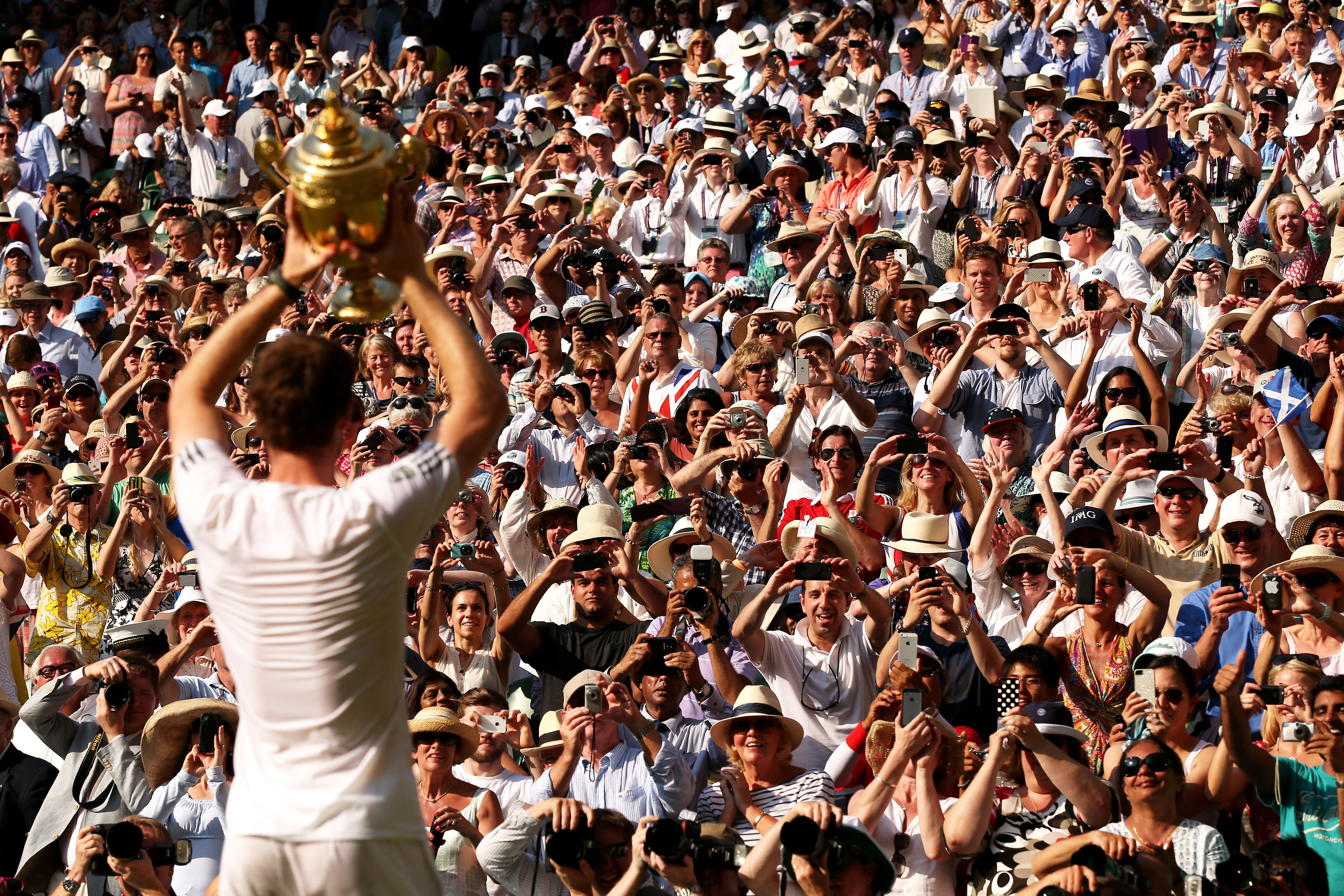 Andy Murray holding the Gentlemen's Singles Trophy aloft to a crowd of cheering fans taking photographs.