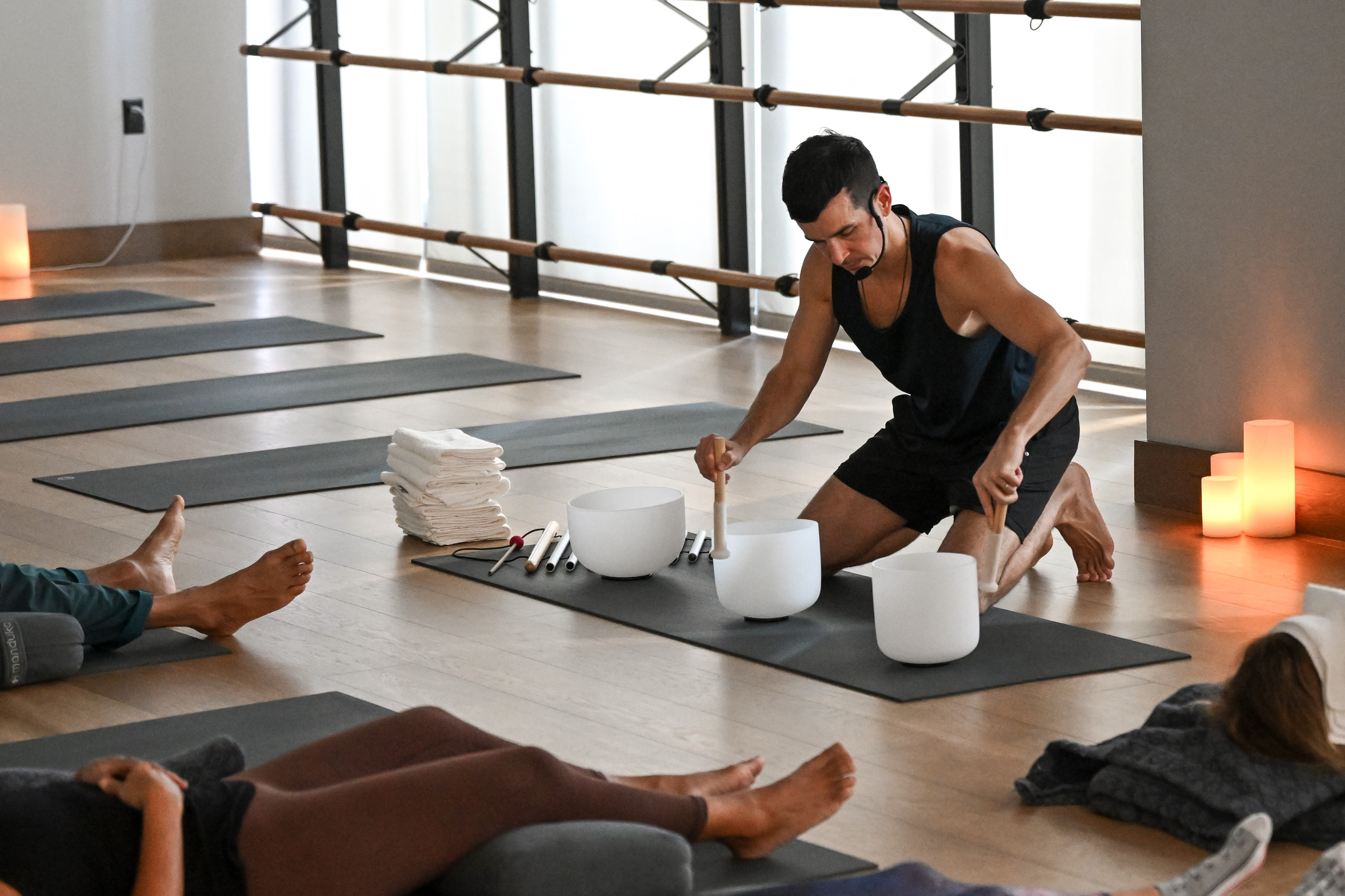 A man leads a sonic meditation class, kneeling to play crystal singing bowls while participants relax on yoga mats.