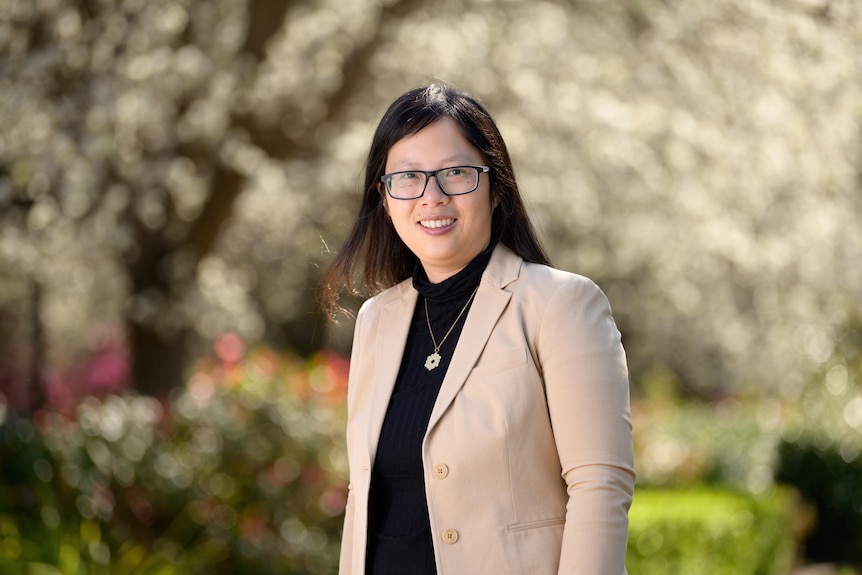 an asian woman with glasses and pink blazer in front of blurred outside background.