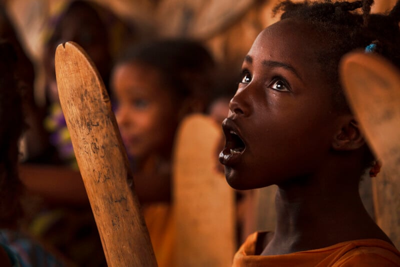 A young girl with braided hair holds a wooden board, looking upward with her mouth open, possibly reciting or singing, in a classroom setting with other children in the background.