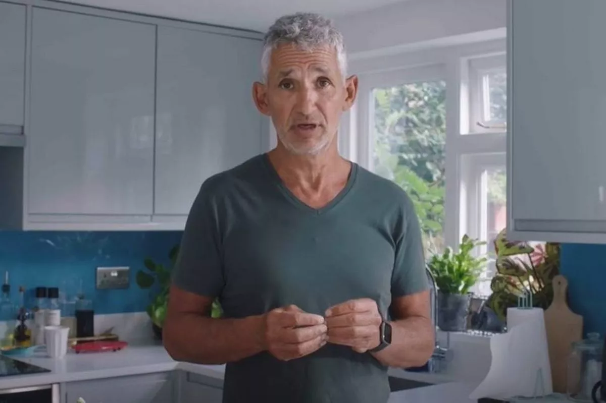 An individual in a kitchen, wearing a gray V-neck t-shirt, standing and gesturing with their hands, with kitchen cabinets, a window, and various kitchen items in the background.