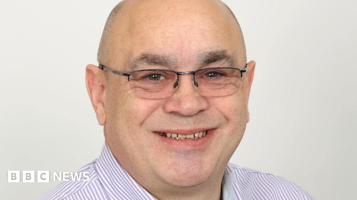 A woman wearing a blue hoodie and a purple jacket with her face buried in her hands, standing in an alleyway.