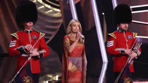 Getty Images Jenkins sings into a microphone, with Beefeaters on either side. She wears a red, blue and white dress which looks like an abstract union jack.