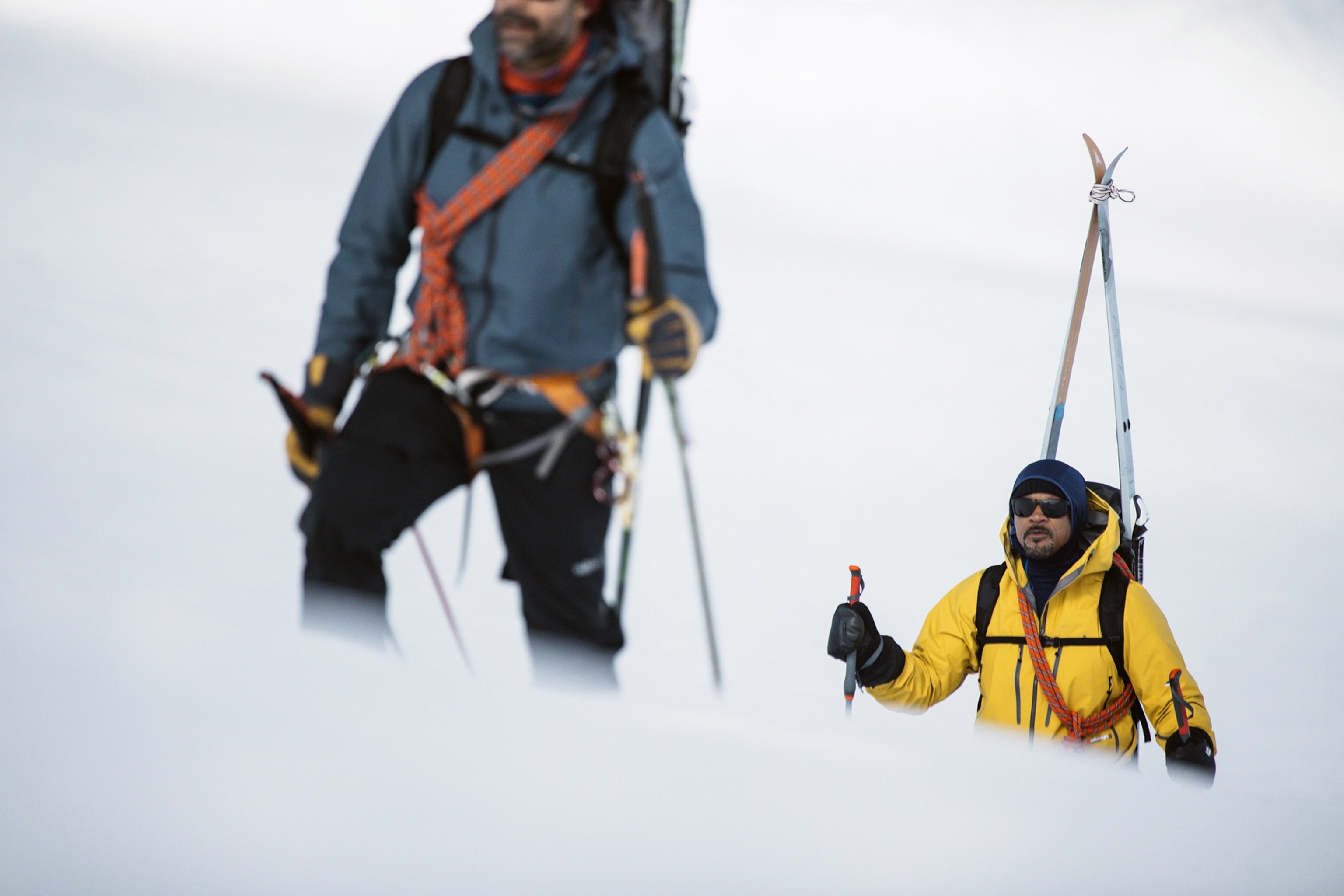 Two climbers, one in foreground, one in background, trek uphill in snow with skis on their backs.