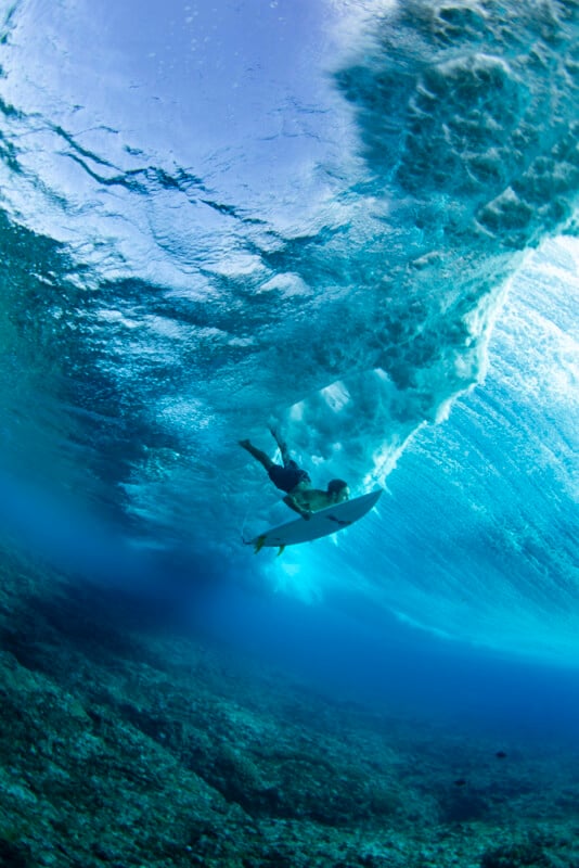 Underwater view of a surfer duck diving beneath a powerful ocean wave, with sunlight filtering through the clear blue water and the rocky seabed visible below.