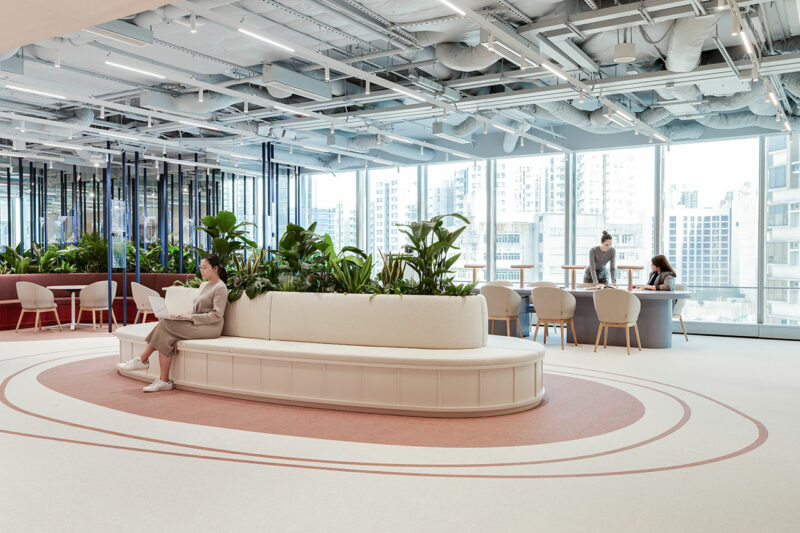 Modern open office with large windows, indoor plants, a curved white sofa, and people working at tables and sitting on the sofa.