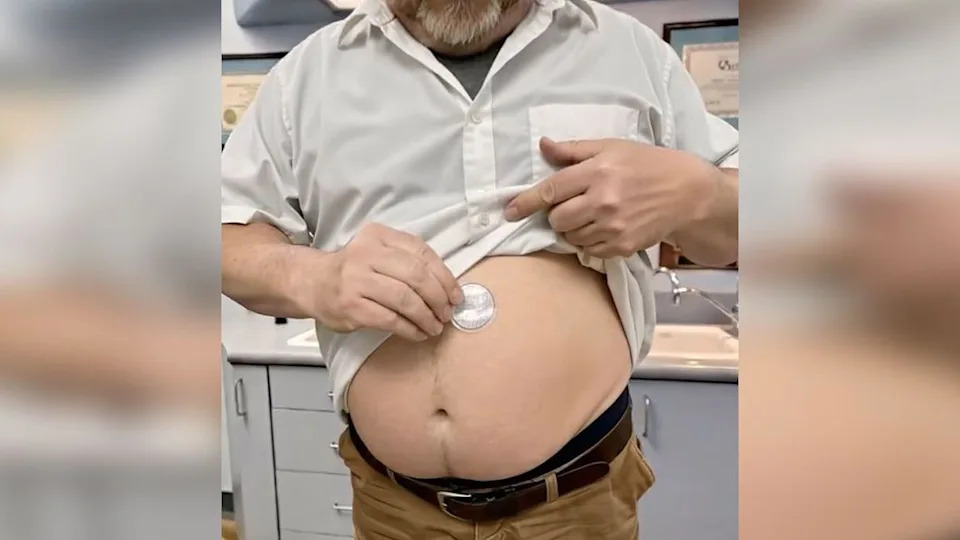 A screen grab from one of the videos which shows a middle aged man lifting a white shirt to reveal a large stomach, whereupon he places a small white patch. The man appears to be in a clinical doctors setting 