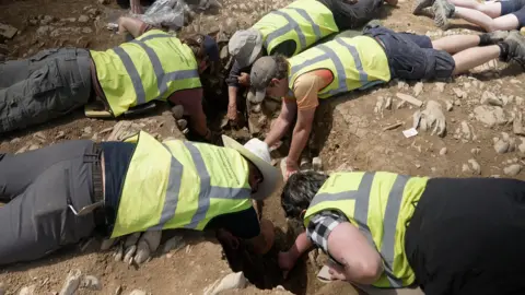 Kevin Church/BBC News Five archaeologists lying face down on the ground clustered around a grave. They are wearing bright yellow high-visibility vests and reaching down into the grave with their hands and tools. 