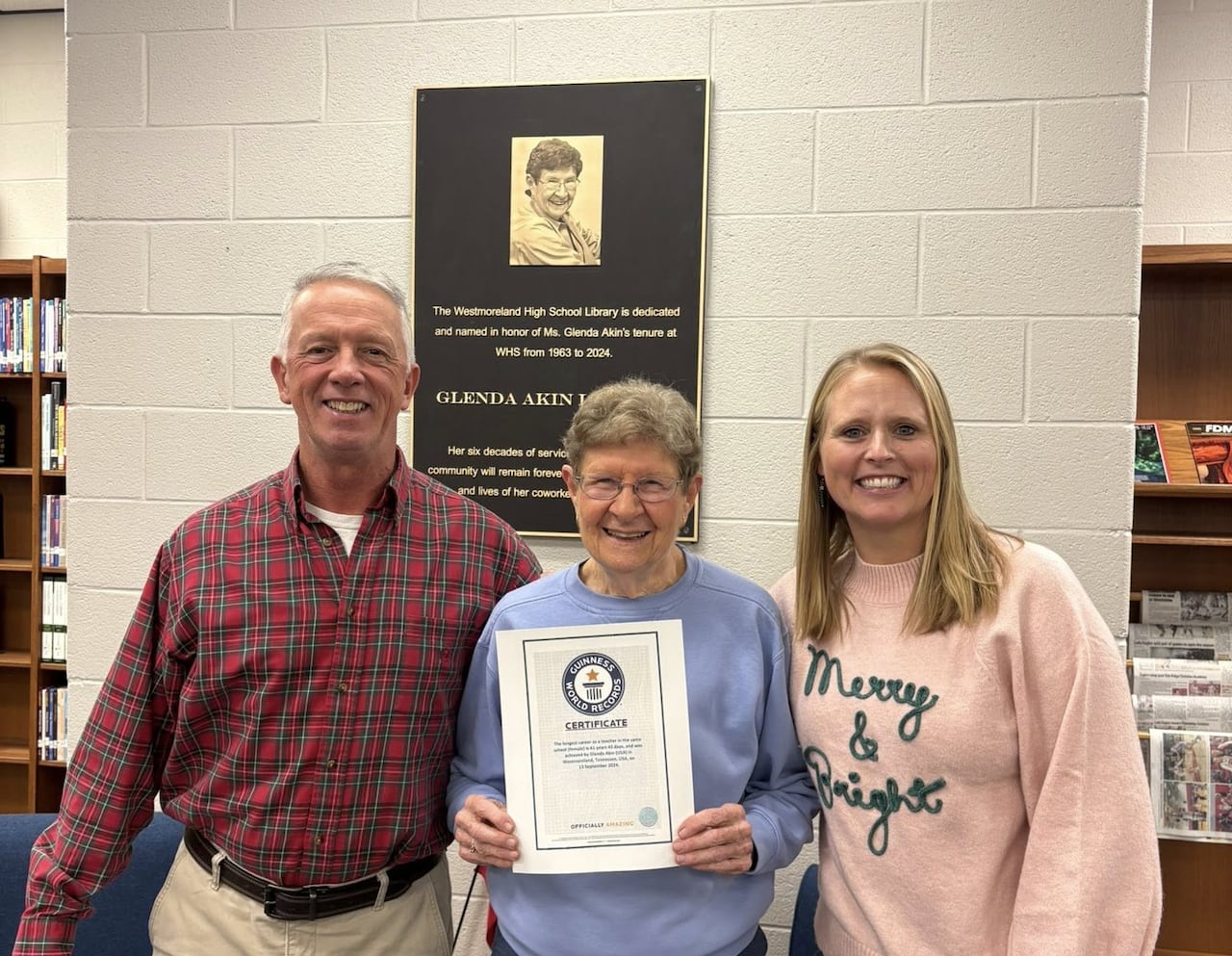 A smiling woman with gray hair stands in a library holding up a Guinness World Record, flanked between a man and a woman. Behind them is a plaque with a picture of the same woman, but younger, and the words: "Glenda Akin Library"