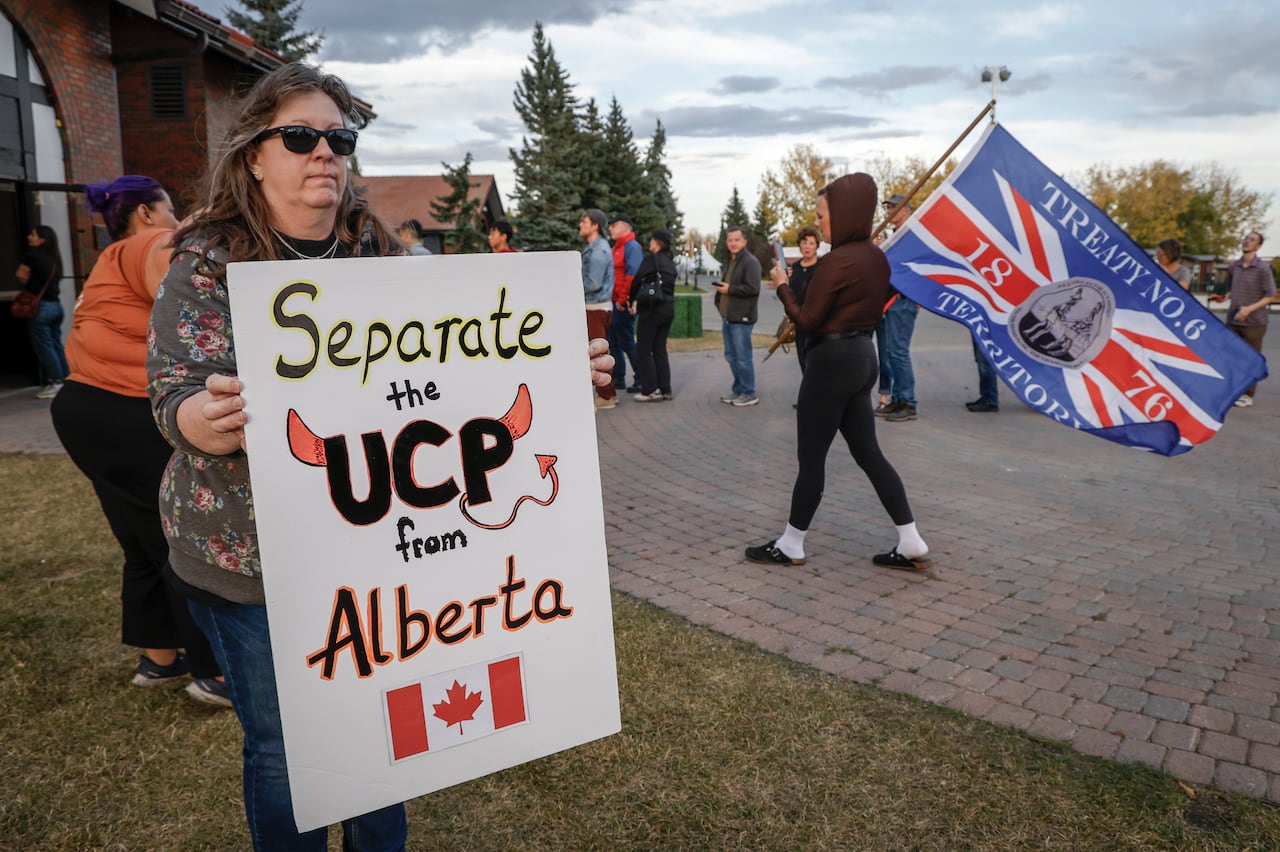 A woman holds a protest sign that says "separate the UCP from Alberta" as people line up in front of a door behind her