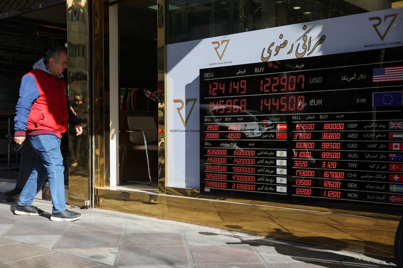 A man in jeans and a jacket walks past the door of a building that displays currency rates on a sign in the window.
