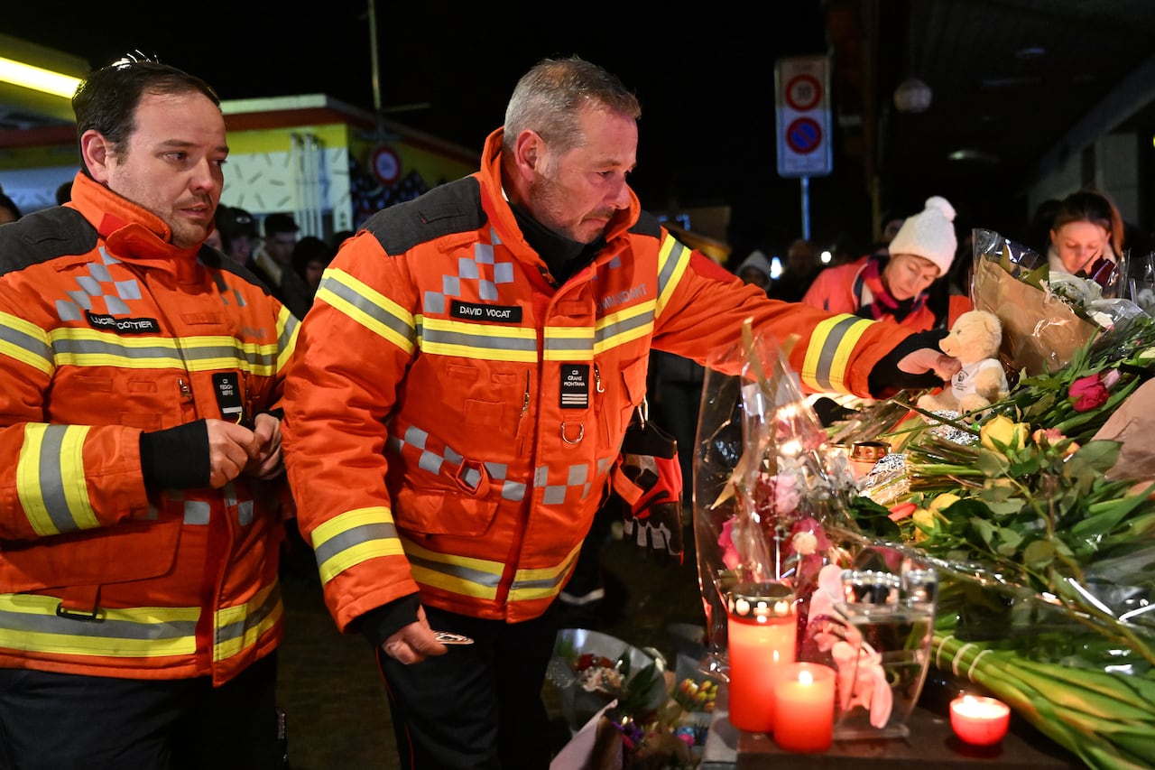 Two men in bright orange firefighter jackets stand beside a makeshift memorial of lit candles and flowers. One of the men places a small teddy bear on the memorial.