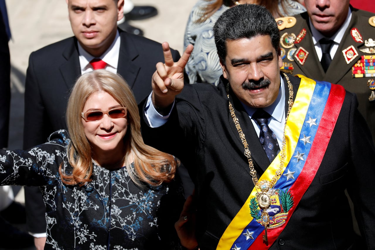 Image of Venezuelan president Nicolas Maduro, dressed in suit and a Venezuelan flag sash across his body and holding up a peace sign, and his wife in sunglasses