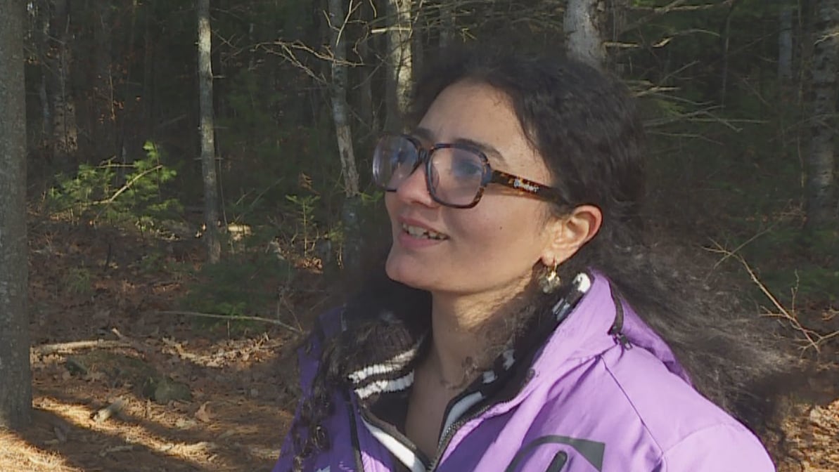 A woman wearing a purple jacket and glasses with black curly hair talks to a reporter