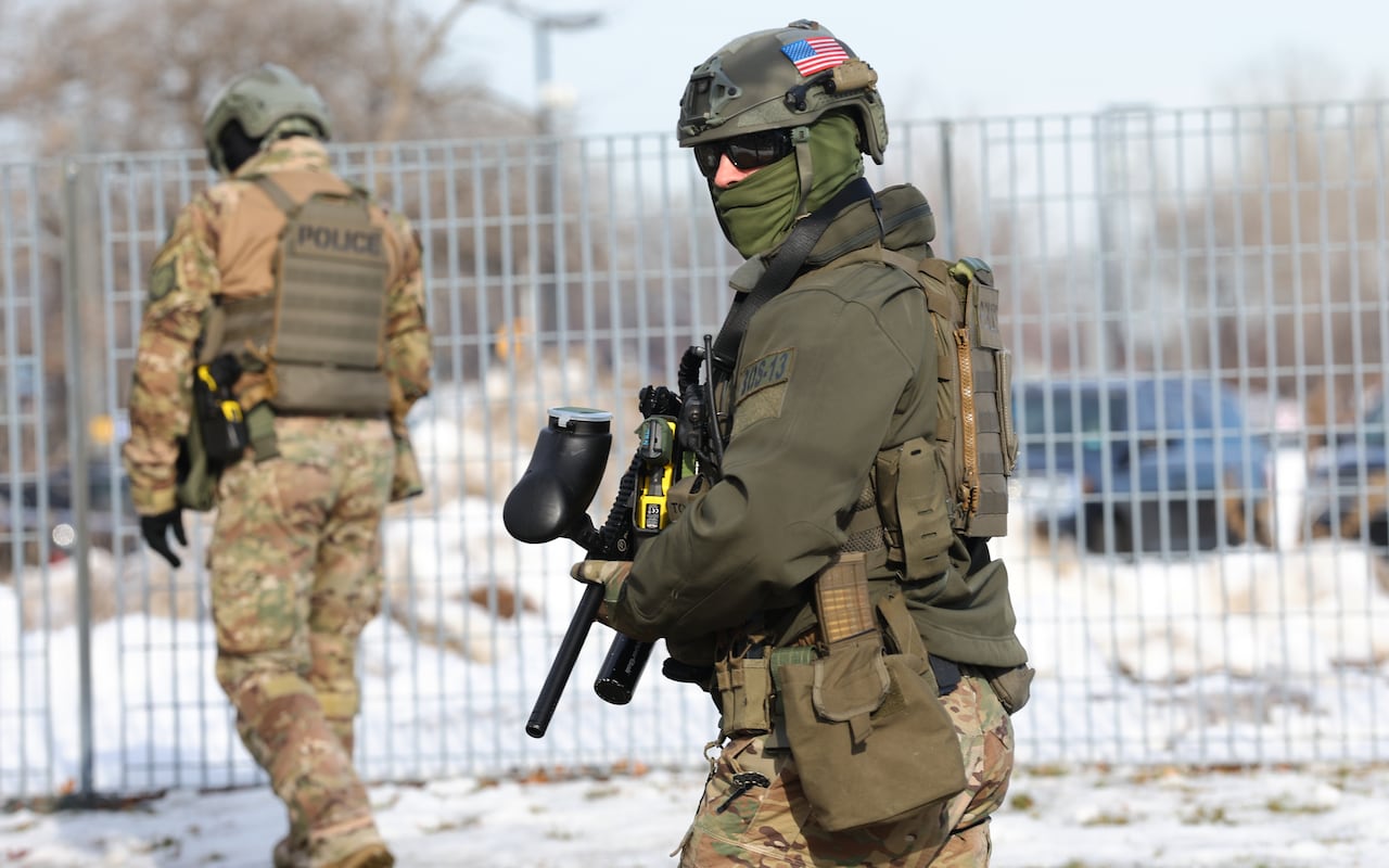 A masked federal agent holding a weapon is seen against a backdrop of a metal fence. 