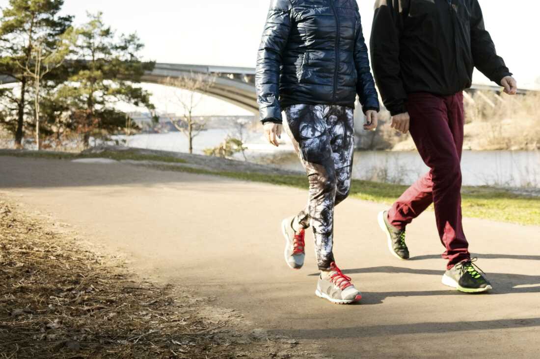 Two people wearing sneakers and photographed from the neck down walk on a paved path that runs along a body of water. A bridge is in the background.