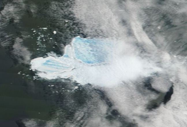 A grainy satellite image of an blue and white iceberg breaking apart on the dark water, with white clouds above. 