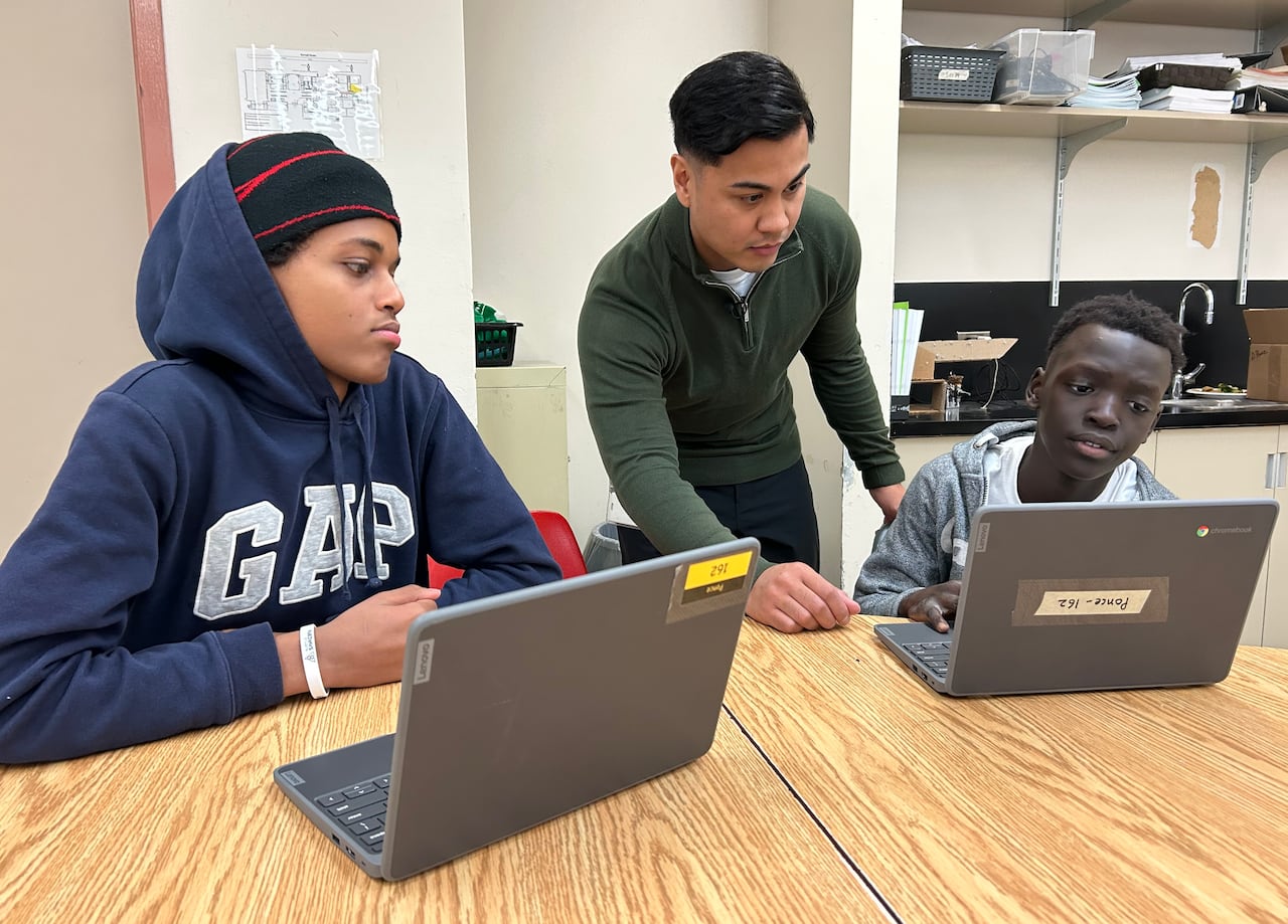 Teacher wearing green stands in the middle of two students sitting at a desk with laptops. 