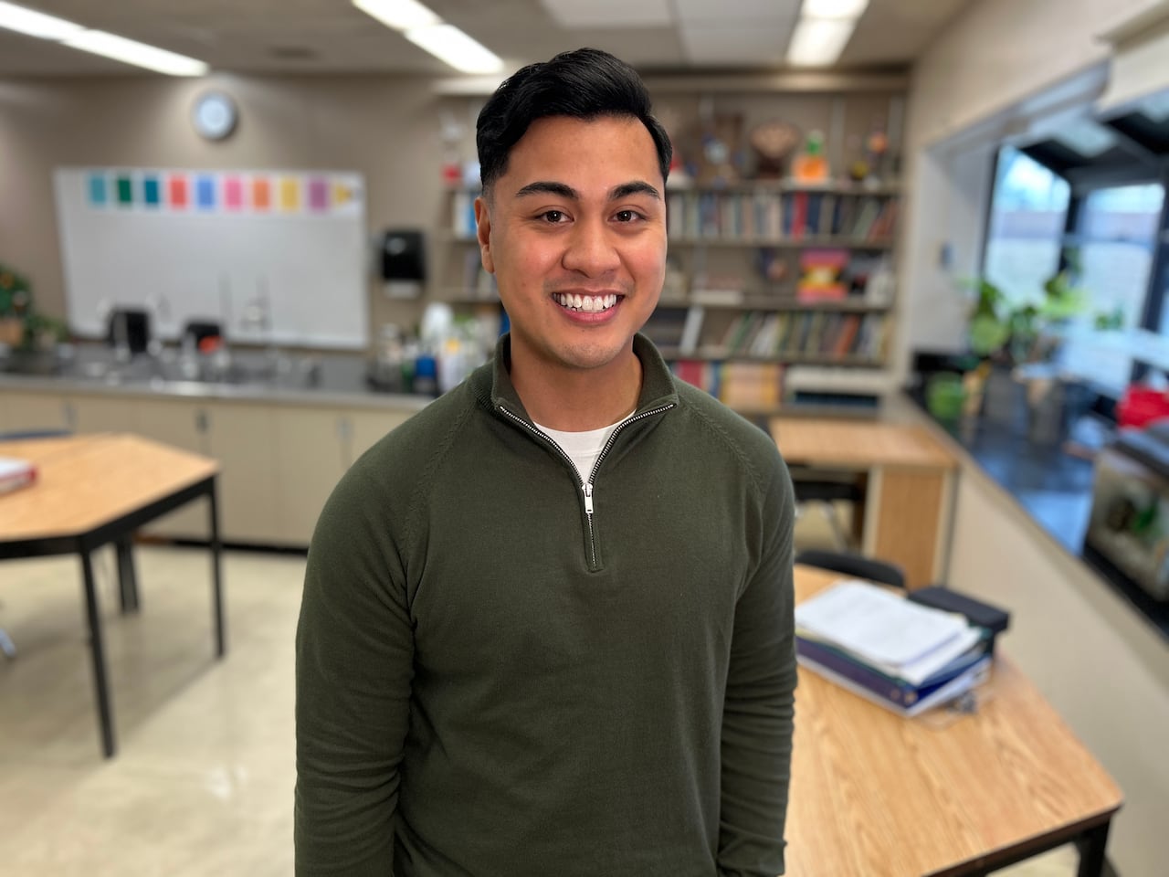 Teacher with brown hair and wearing green sweater stands in an empty classroom.