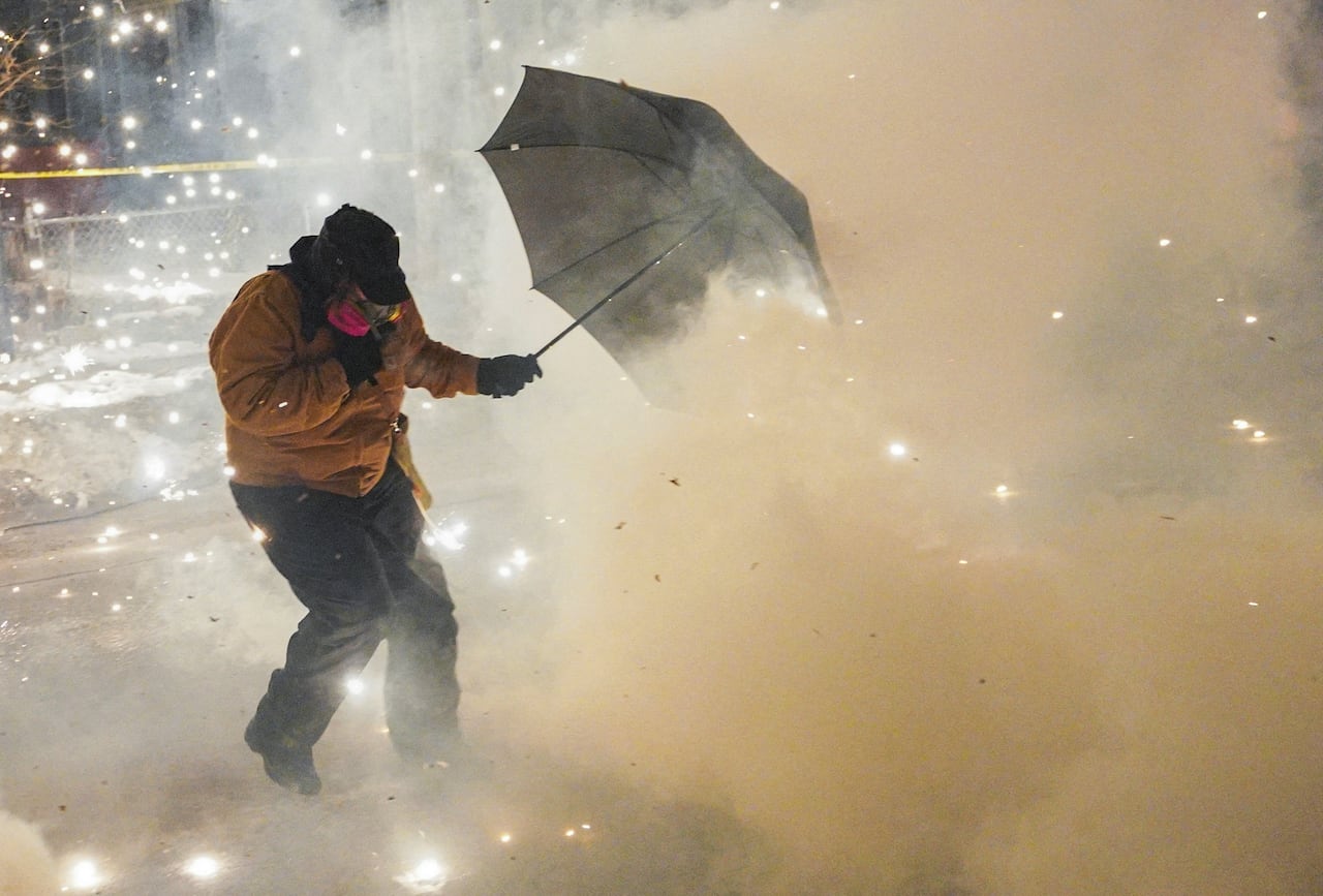 A person wearing winter clothing and carrying an umbrella appears to shield themselves from pepper balls causing smoke.
