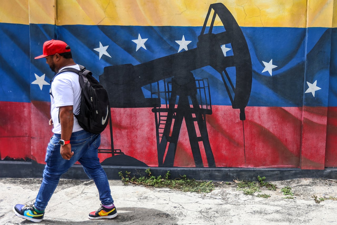 A man in a backpack walking past a mural showing an image of an oil pumpjack and the Venezuela flag