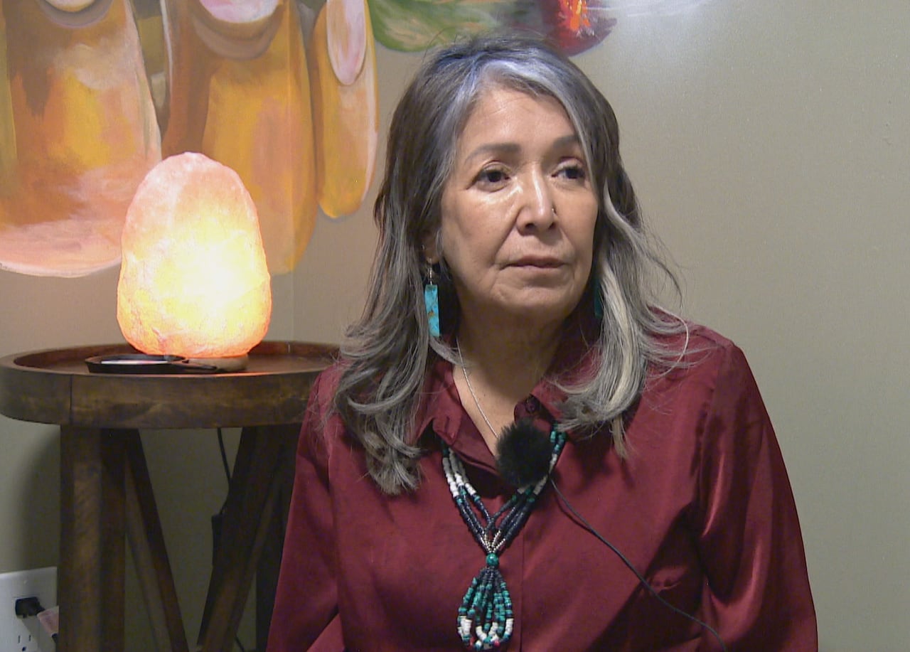 A woman with long gray hair wearing a burgundy shirt and a large beaded blue necklace. 