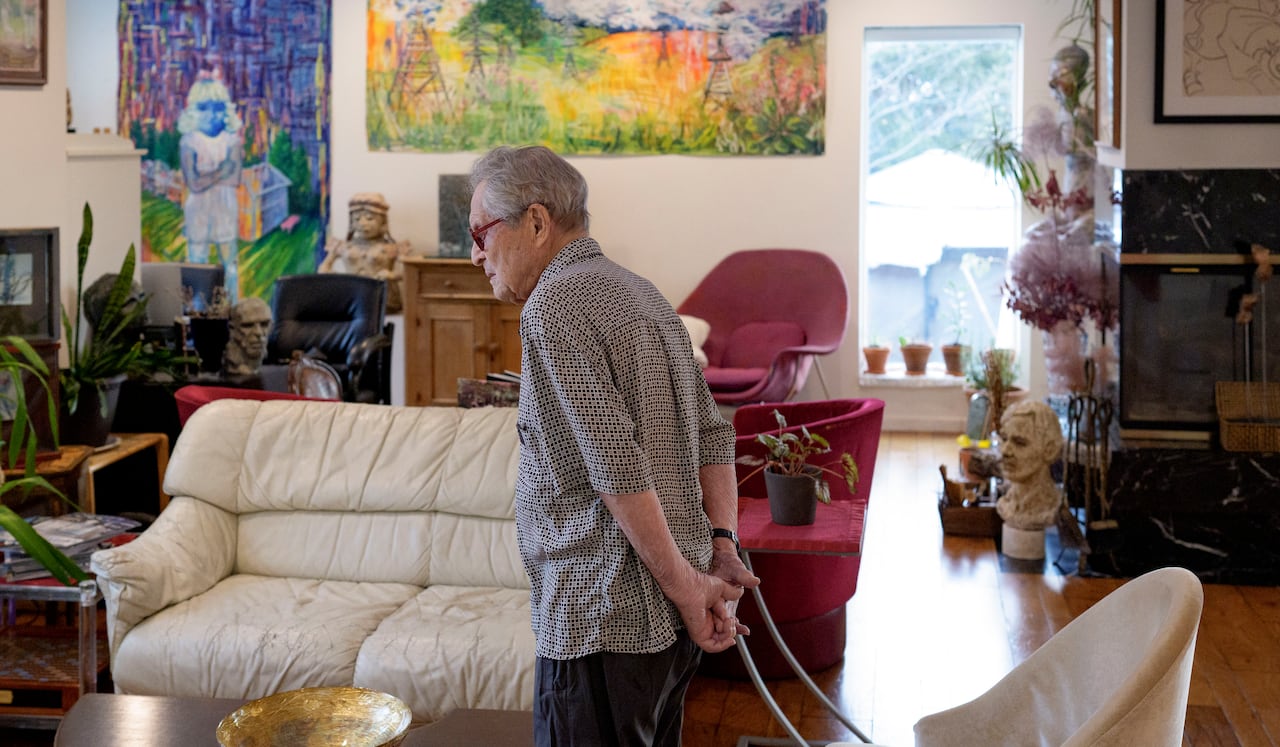 A man with grey hair stands with his hands behind his back in a living room filled with art and plants. 
