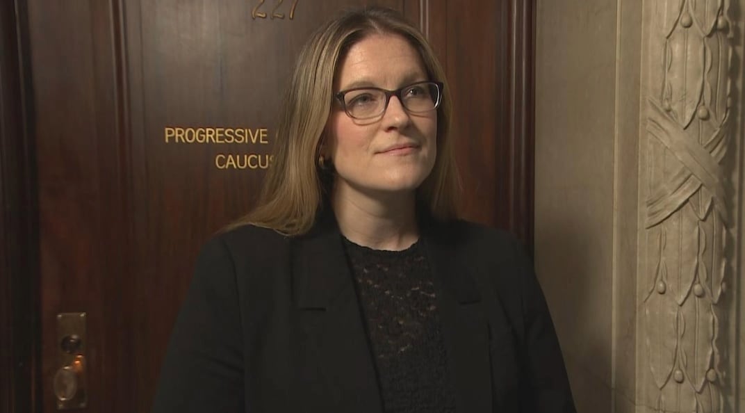 A woman with long hair wearing glasses, a black top and blazer stands in front of a door that says "Conservative Caucus" on it. 