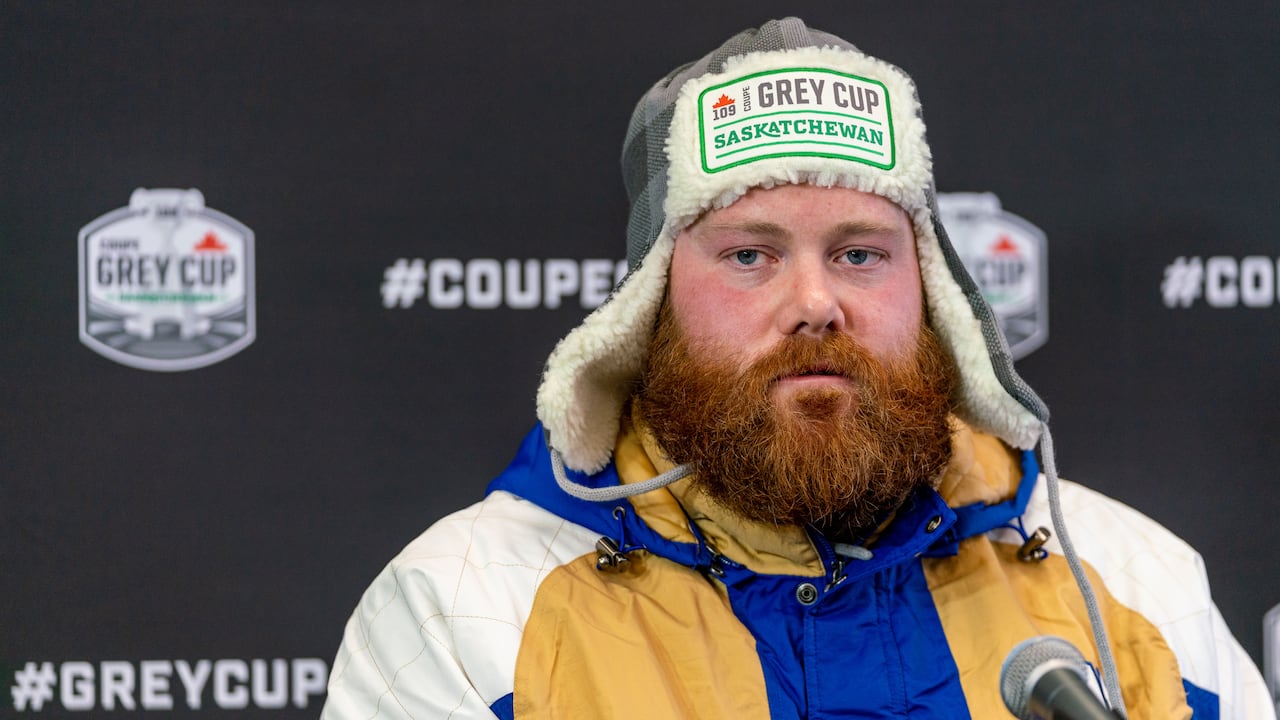 Man with red beard wears a blue, yellow and white jacket and a fleece hat that says "Grey Cup Saskatchewan" on it