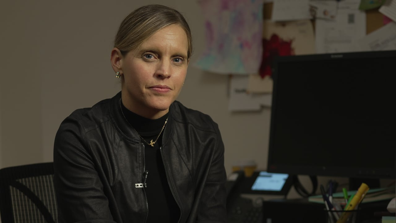 A woman with blonde hair pulled back in a ponytail wears a black jacket and shirt while sitting at a desk and looking into the camera. 