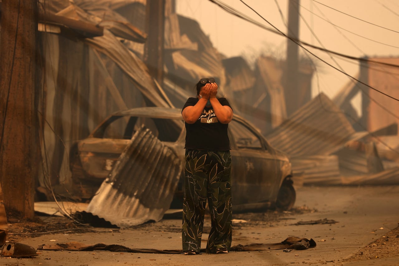 A person covers their face with their hands as they stand amid the charred remains of buildings and vehicles.
