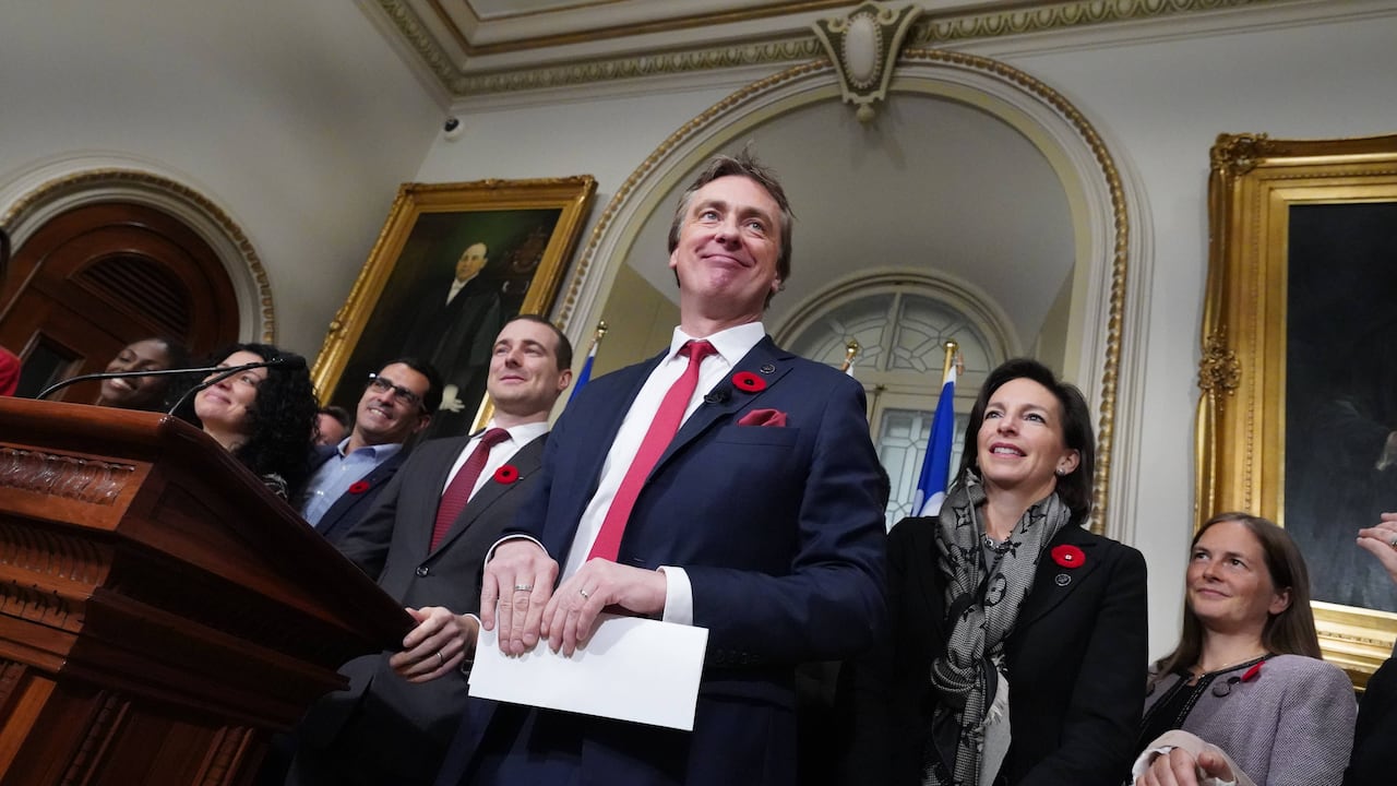 An angled shot of a guy in a suit, surrounded by men and women with an ornate background.