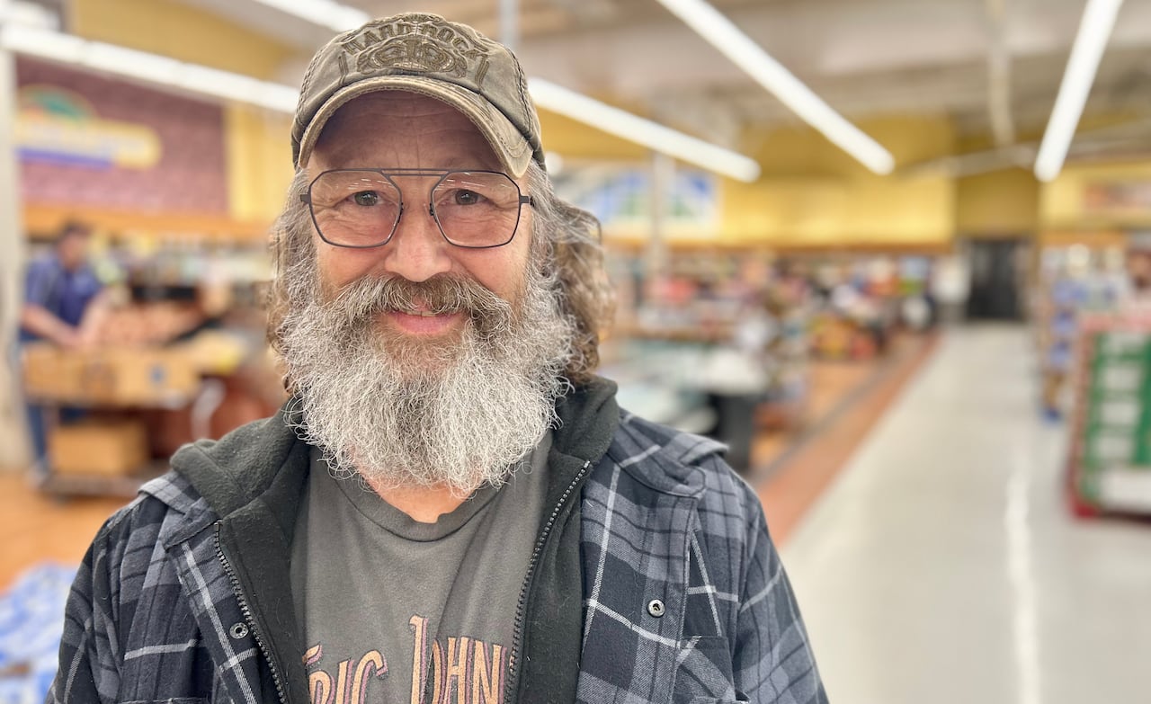 A man with a brown ball cap, glasses and a greyish beard smiles at the camera while standing in a grocery store. 