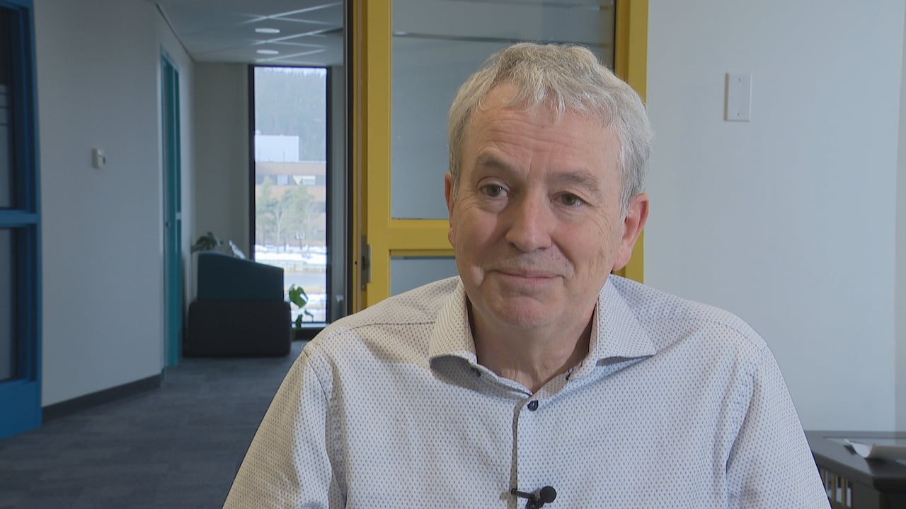 A middle-aged man in a white patterned shirt sits in a chair in an office 