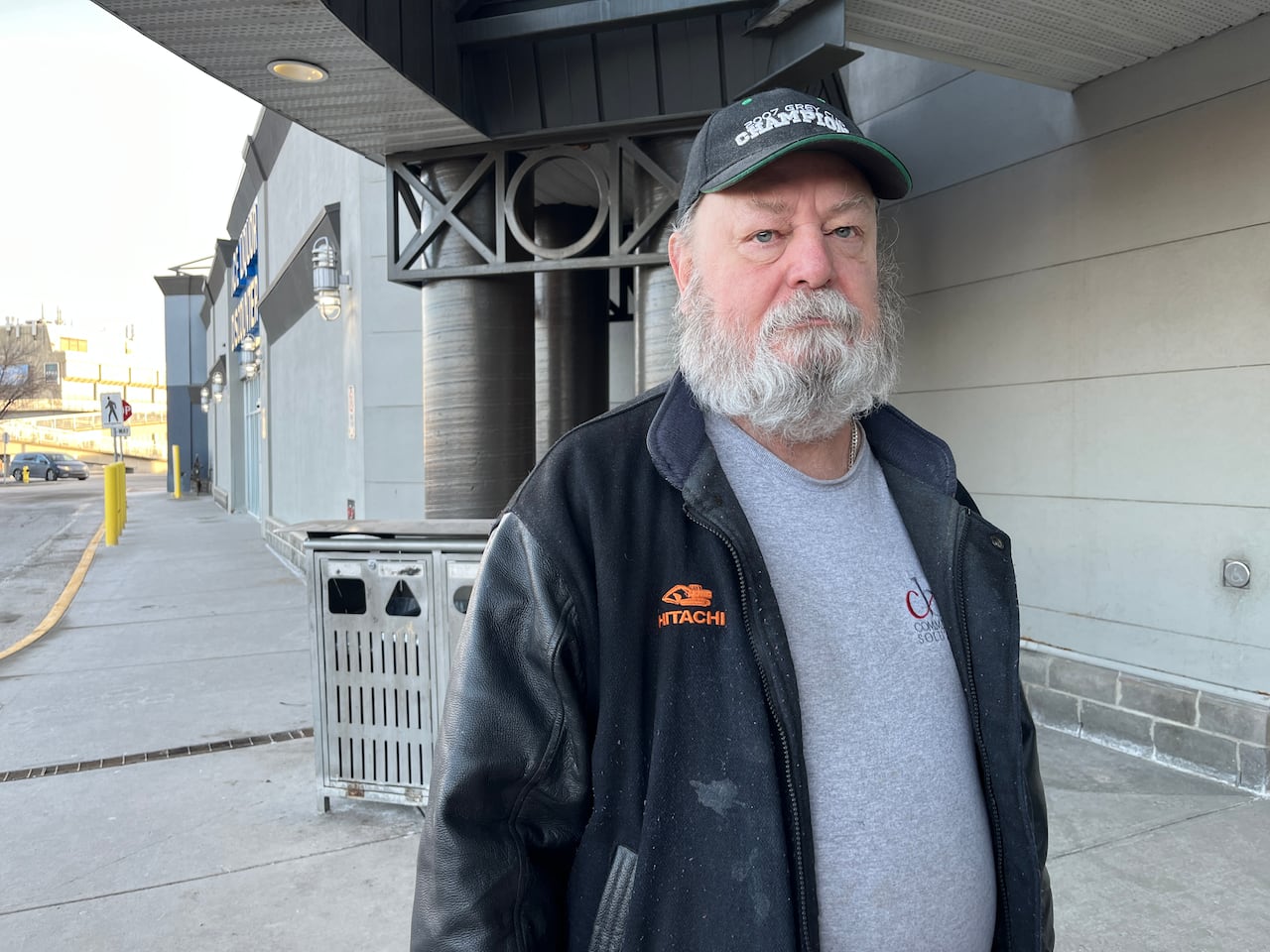 A man with a grey beard and a baseball cap is shown outside a shopping centre.