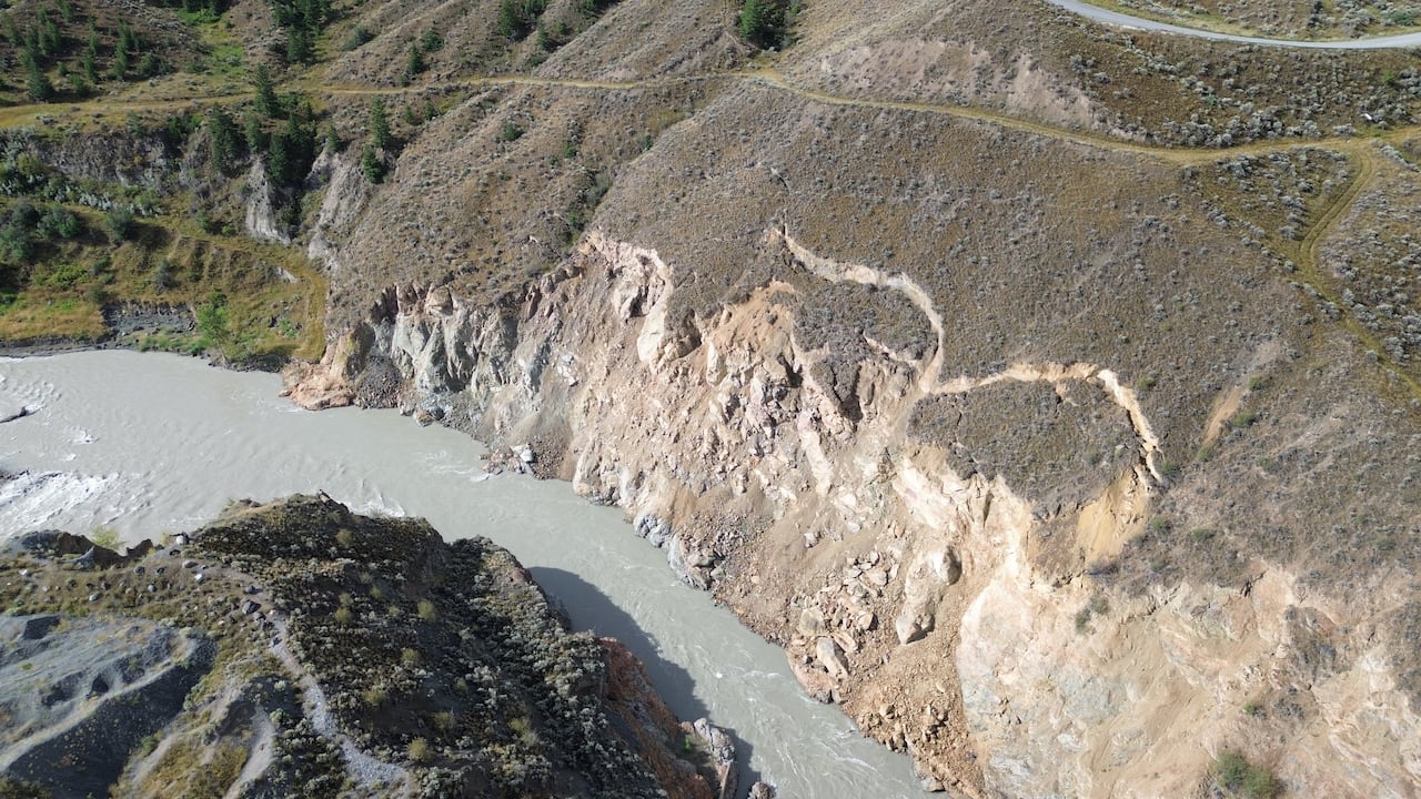 A hillside above a grey river is seen with cracks in it.