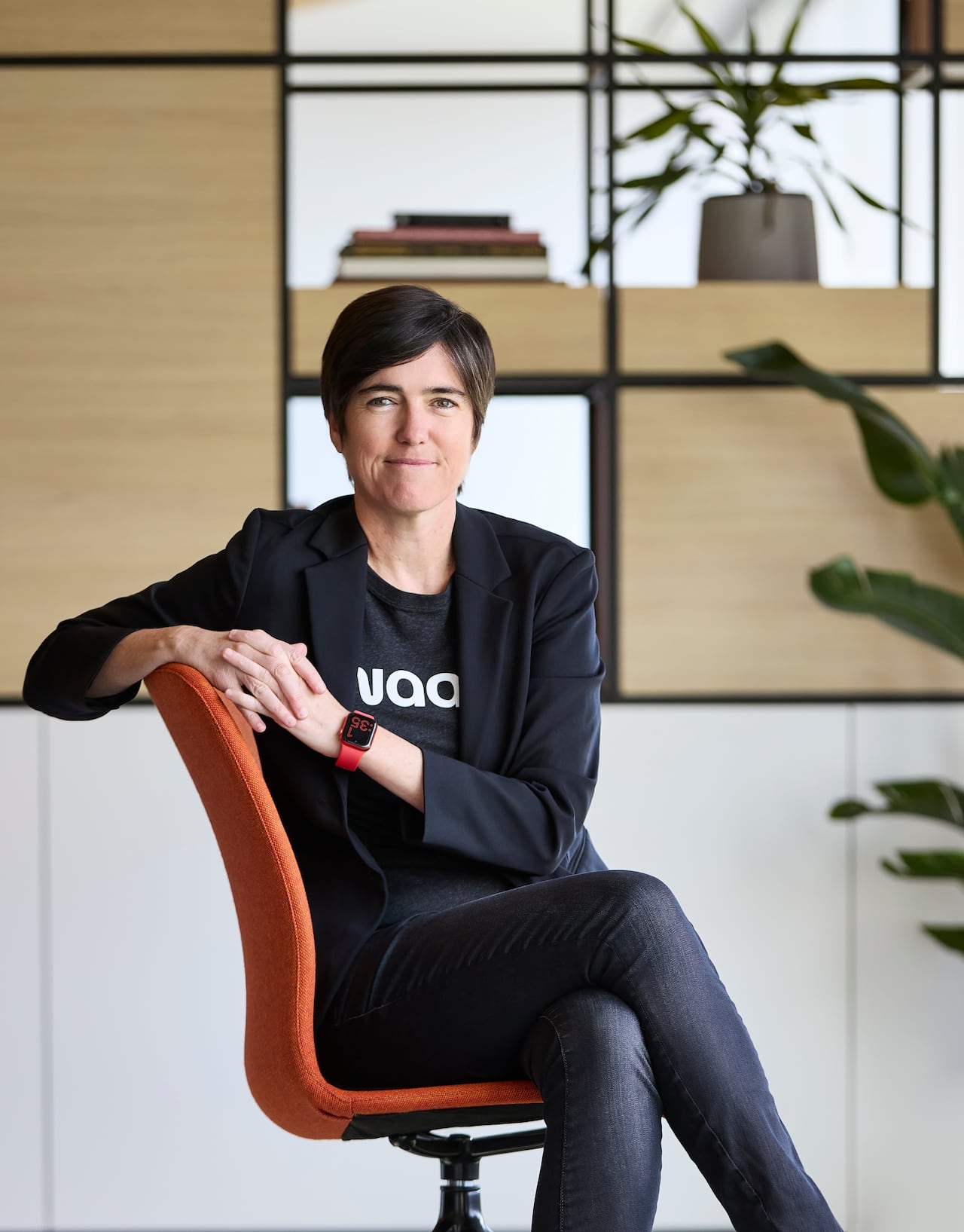 A woman with short brown hair sits on an orange chair and smiles at the camera