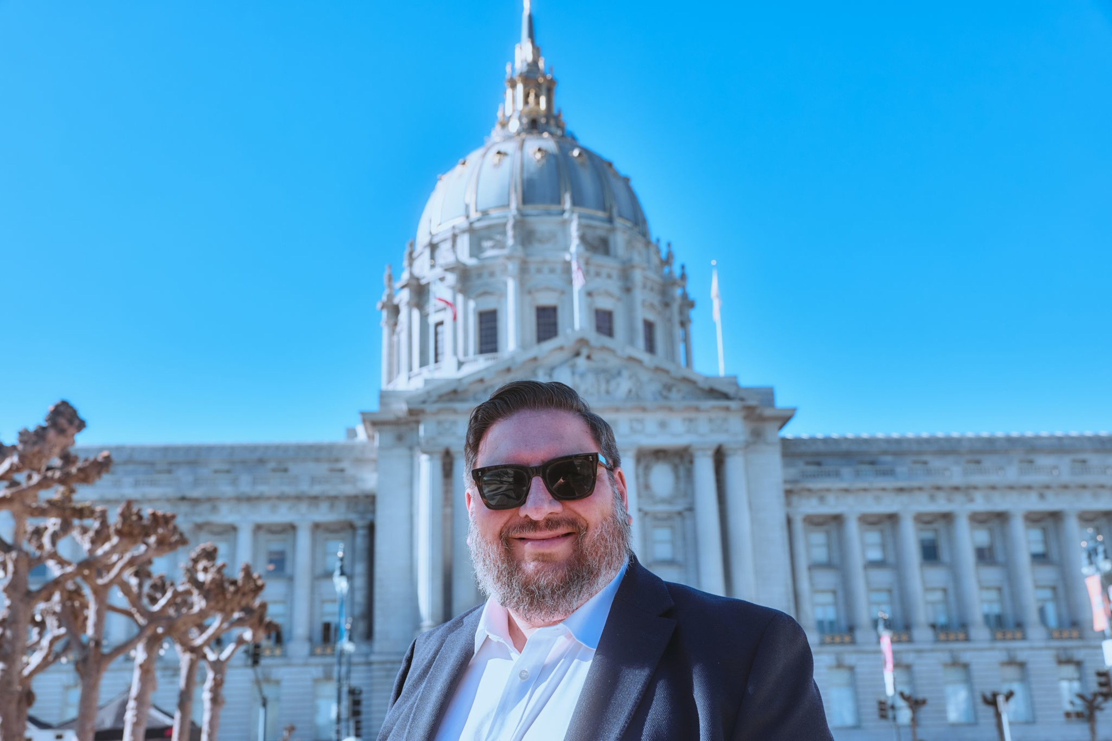A bearded man in sunglasses and a suit smiles in front of a large domed government building under a clear blue sky.