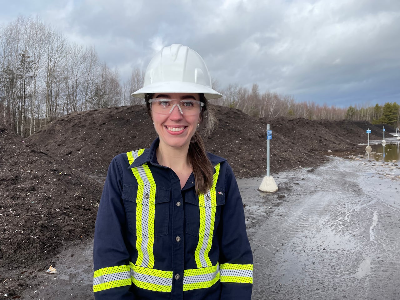 A woman in a reflective vest, white hard hat and protective glasses smiles in front of piles of compost that have almost finished maturing.