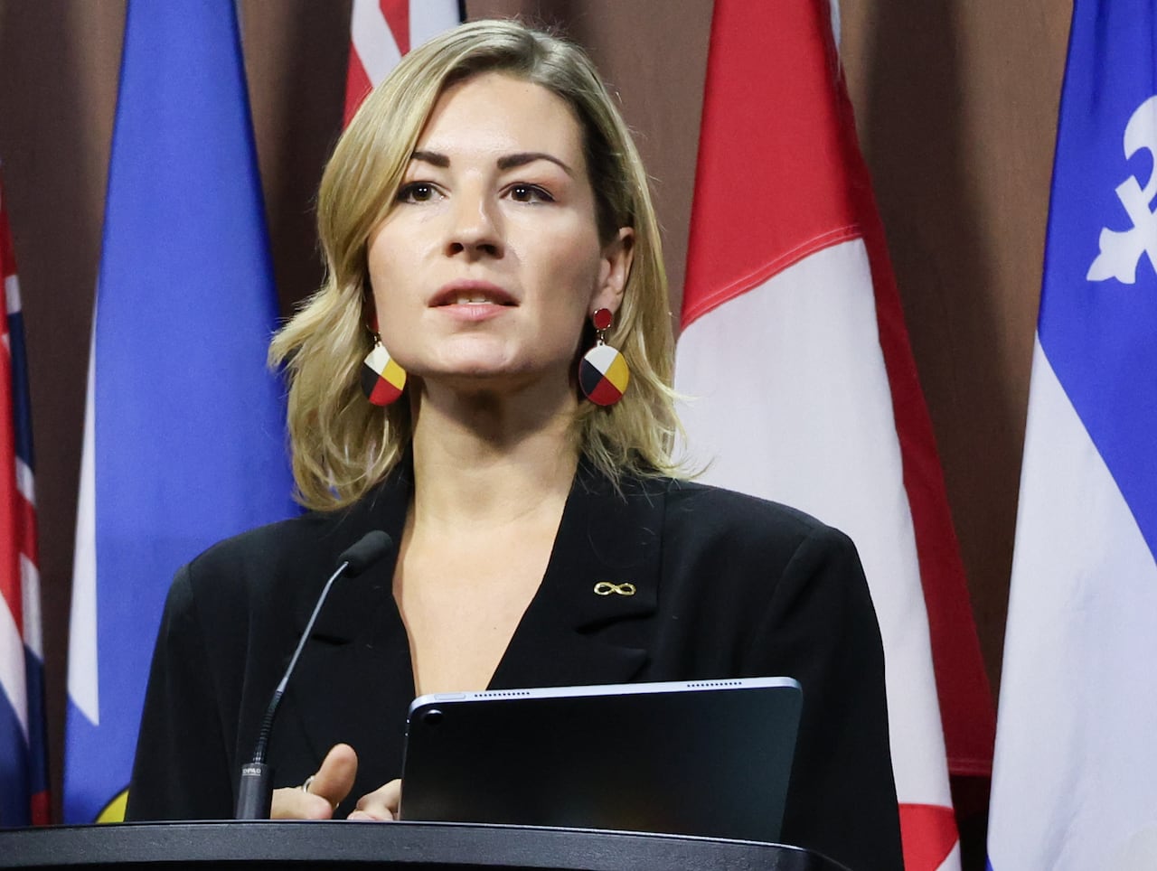 Amy Lamb wearing a black blazer with a Metis nation pin and medicine wheel earrings stands at a podium with provincial flags behind her
