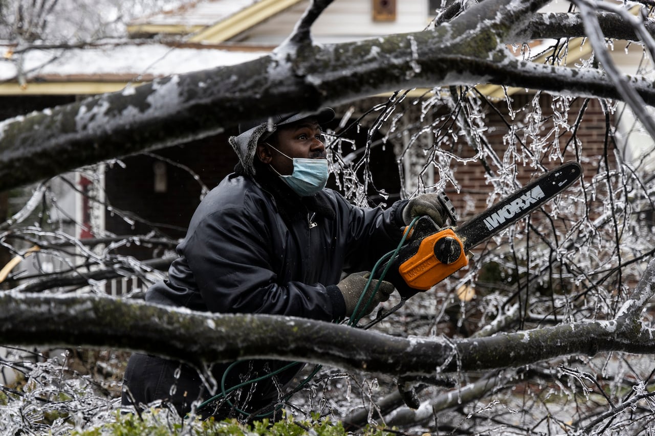 A man wearing a black hooded coat and a light green medical mask on his face, holds a chainsaw as he walks between the ice-covered branches of a large fallen tree on a residential street.