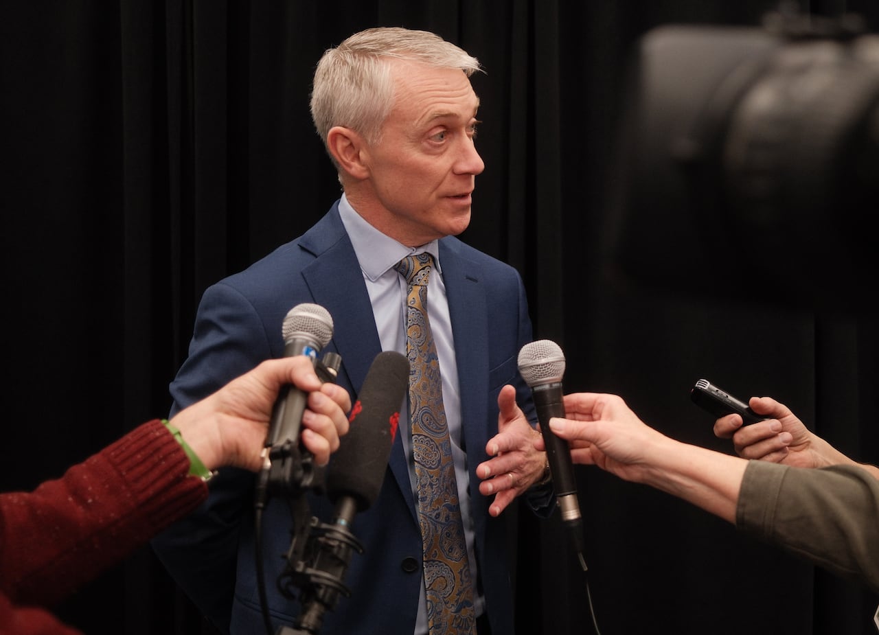A man in a suit and tie stands in front of outstreatched arms holding microphones.