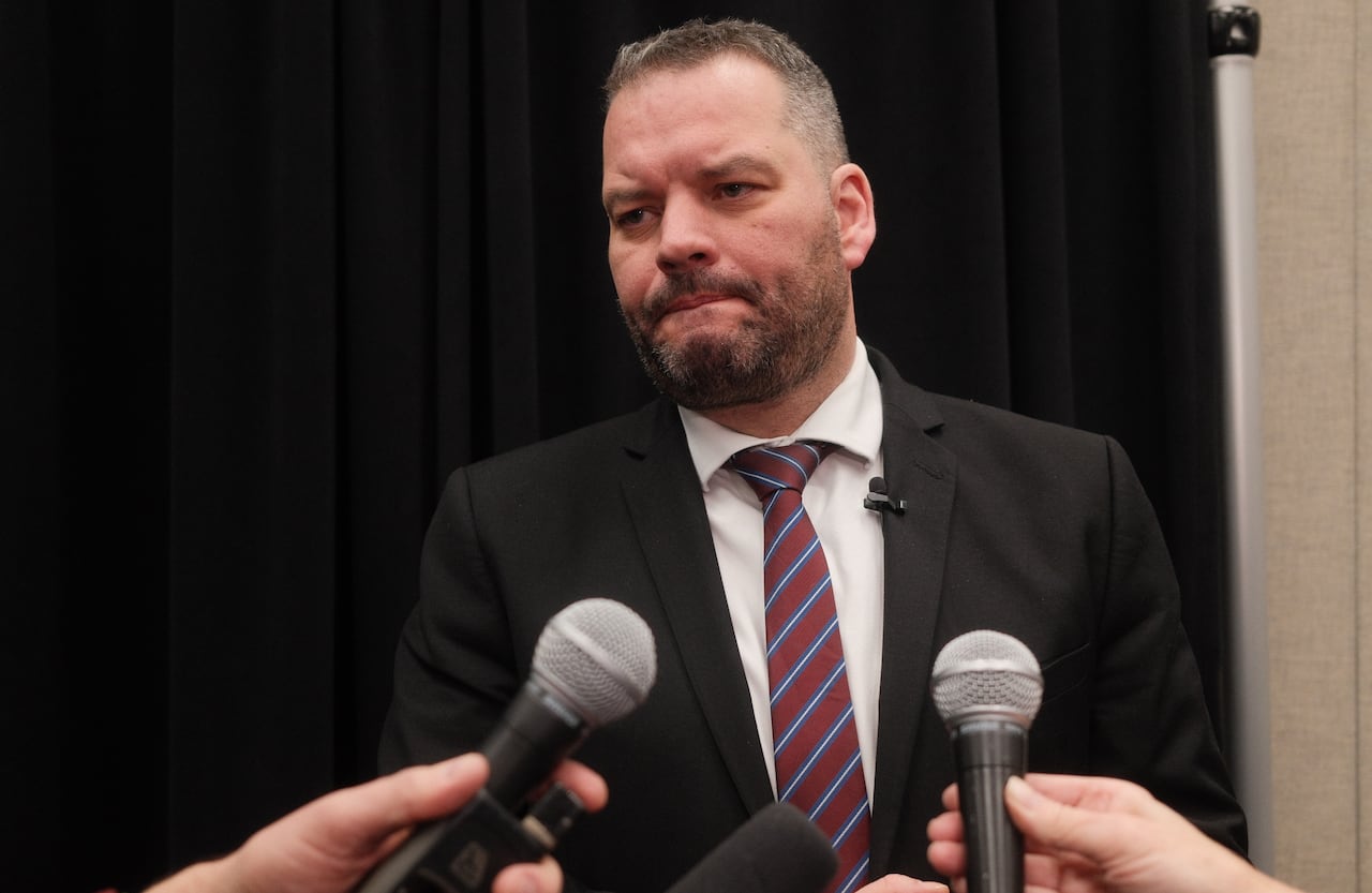 A man in a suit and tie stands in front of outstretched arms holding microphones.