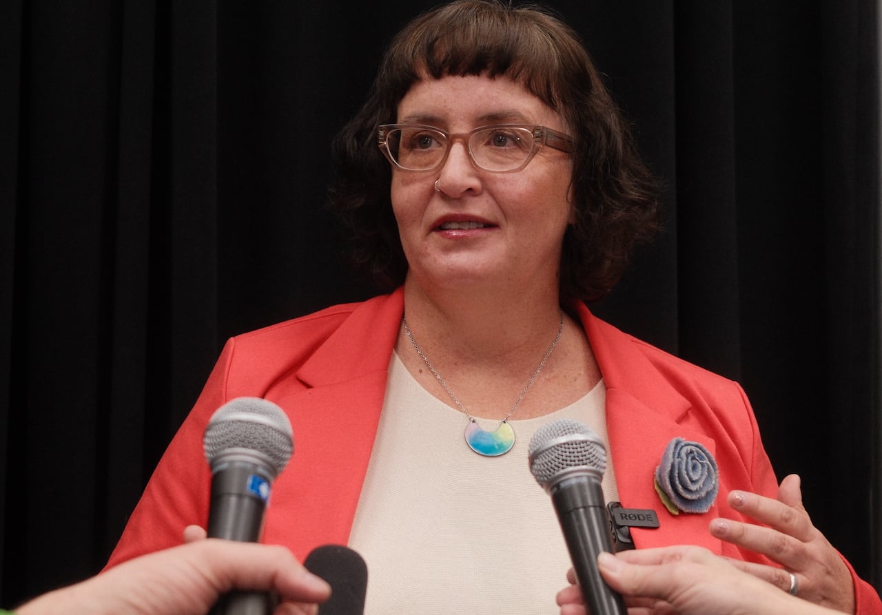 A person with glasses and a peach sport jacket stands in front of microphones.