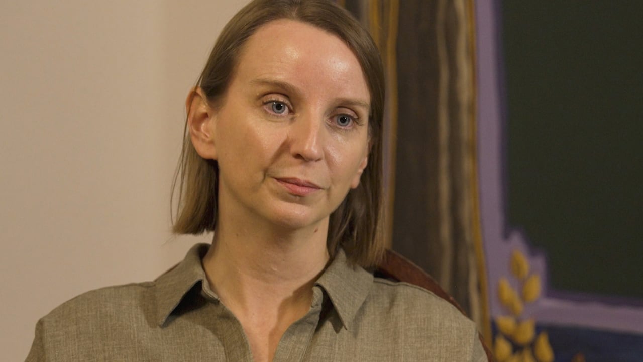 A woman looks pensive in a room while wearing a grey shirt.