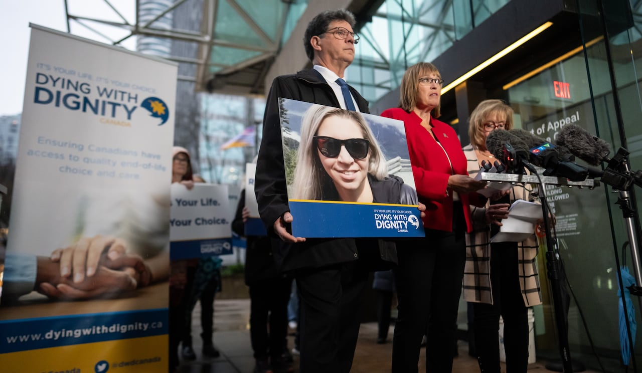 A group of people stand in front of a banner marked 'Dying with Dignity', holding a photo of a blond woman.