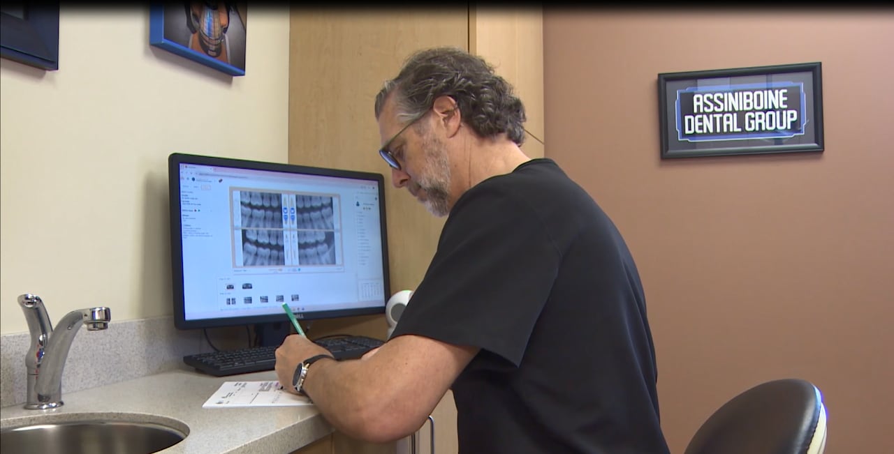 Dr. Scott Leckie wearing black scrubs sits at a desk with his computer and fills out paperwork by hand at his clinic in Winnipeg.
